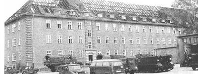 Ein großes, abgebranntes Gebäude mit beschädigtem Dach und parkenden Fahrzeugen davor nach einem Brand.