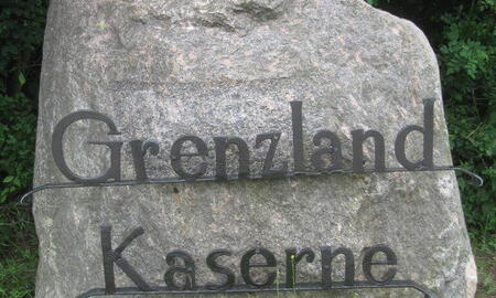 Ein großer Stein mit der Aufschrift "Grenzland Kaserne" steht vor einer Kulisse aus grünen Büschen.