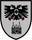 Ein Wappen zeigt einen schwarzen Doppeladler mit grünem Herzschild über einem stilisierten Gebäude auf grauem Hintergrund.