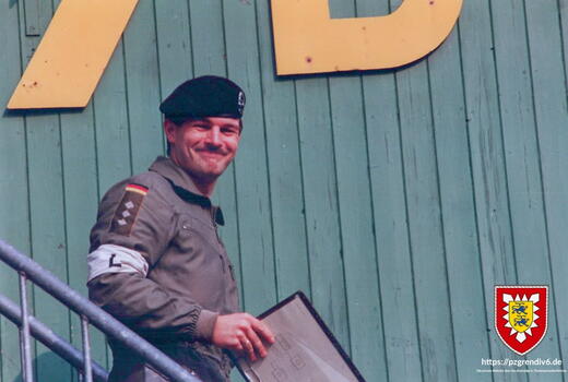 Ein Soldat in Uniform und Barett steht lächelnd auf einer Treppe vor einer Holzwand und hält eine Mappe.
