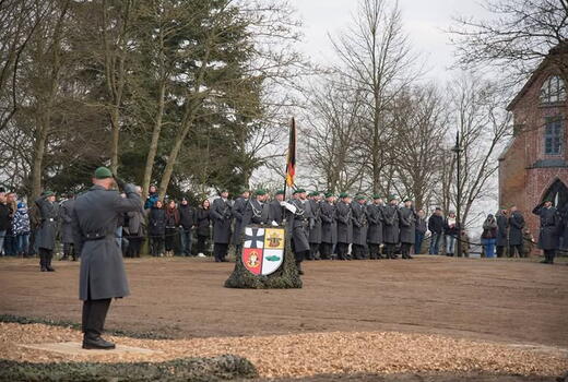 Eine Militärzeremonie mit Soldaten in grauen Mänteln und grünen Baretten, die neben einem Wappen-Standarte salutieren.