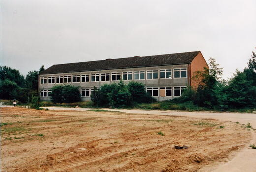 Ein graues, zweistöckiges Gebäude der Bismarck-Kaserne von 1996 steht umgeben von Bäumen und einer unbefestigten Fläche.