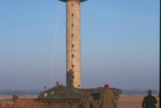 Soldaten in Uniform stehen vor einem hohen Wachturm auf einem Feld, neben einem Panzerfahrzeug bei klarem Himmel.