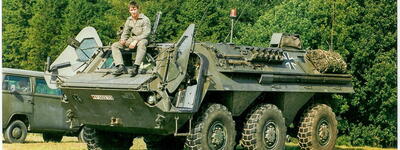 Ein Mann sitzt auf einem gepanzerten Militärfahrzeug, umgeben von grünen Bäumen, neben einem olivfarbenen Transporter.