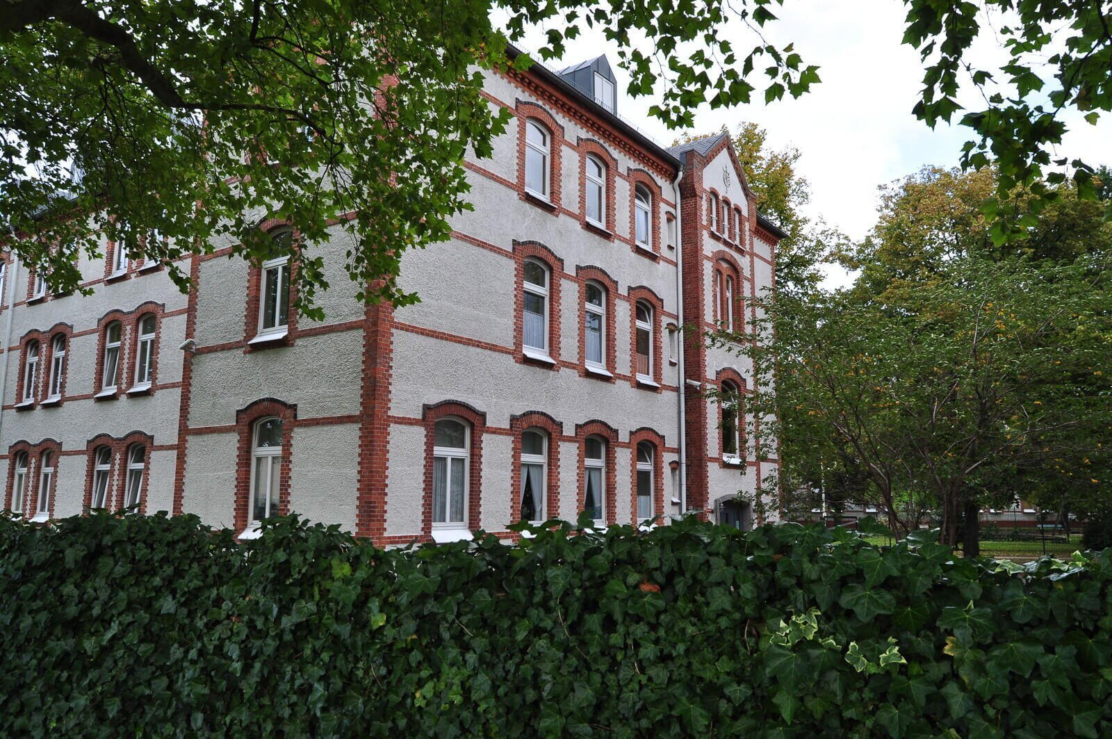 Ein historisches Backsteingebäude mit roten Akzenten steht hinter einer grünen Hecke, umgeben von Bäumen.
