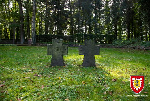 Zwei moosbedeckte Kreuze stehen auf einem gepflegten Rasen in einem ruhigen, von Bäumen umgebenen Friedhof.