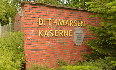 Der Eingang zur Dithmarsen-Kaserne zeigt eine Backsteinmauer mit gelber Schrift und umgebender grüner Vegetation.