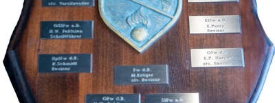 Ein hölzernes Schild mit Metallplatten zeigt die Gründungsmitglieder des Raketenkartellbataillons.