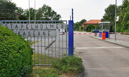 Eingang der Stapelholmer Kaserne mit Schranke und Wachhäuschen, umgeben von grüner Vegetation und einem klaren Himmel.