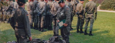 Soldaten in Uniform versammeln sich auf einer Wiese zur Waffenausgabe, während ein Offizier Unterlagen überprüft.