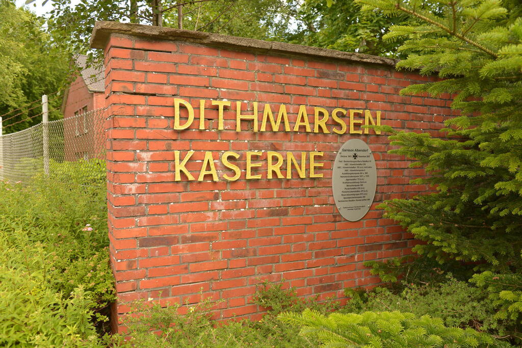 Der Eingang zur Dithmarsen-Kaserne zeigt eine Backsteinmauer mit gelber Schrift und umgebender grüner Vegetation.