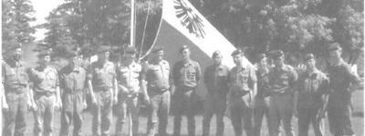 Eine Gruppe von Soldaten steht in Uniform vor einer Fahne mit Adleremblem auf einer Wiese.