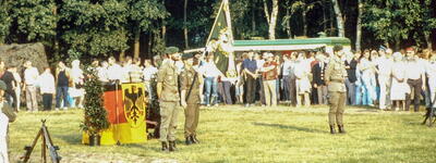 Soldaten in Uniform stehen neben einer deutschen Flagge, während eine Gruppe von Menschen bei einer Herbstveranstaltung zusieht.