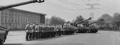 Soldaten stehen in Formation neben Panzern auf einem Übungsplatz mit einem großen Gebäude im Hintergrund.