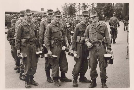 Eine Gruppe von Soldaten in Uniformen steht mit Ausrüstungsgegenständen auf einem offenen Platz zusammen und wirkt entspannt.
