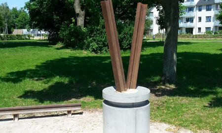 Eine abstrakte Skulptur aus Metall erhebt sich aus einem Betonfundament in einem grünen Park unter Bäumen.