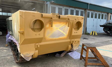 Ein gelber Militärpanzer wird vor einer Werkstatt frisch lackiert, während eine Holzleiter daneben steht.