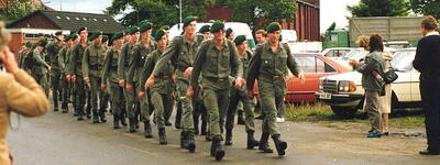 Eine Gruppe Soldaten in grünen Uniformen marschiert auf einer Straße, während Zuschauer am Rand stehen.