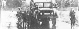 Eine Gruppe von Soldaten steht und sitzt auf einem Militärfahrzeug auf einer nassen Straße.