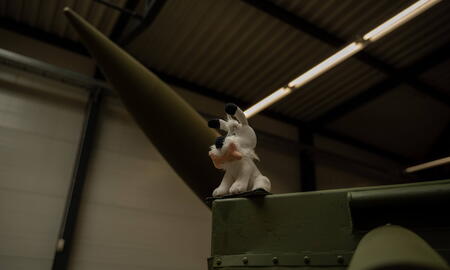 Ein weißer Plüschhund sitzt auf einem grünen Militärfahrzeug in einer Halle mit einem großen Raketenobjekt im Hintergrund.