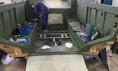 Ein Mechaniker arbeitet in einer Werkstatt an einem großen, grünen Militärfahrzeugrahmen mit Werkzeugen und Ausrüstung.