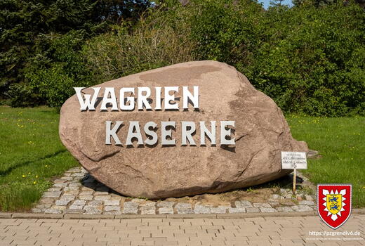 Ein großer Stein mit der Aufschrift "Wagrien Kaserne" steht auf einem gepflasterten Platz, umgeben von grünem Rasen.