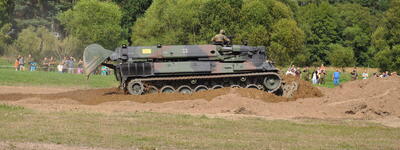 Ein Panzer bewegt sich durch eine sandige Grube, während Zuschauer aus sicherer Entfernung zusehen.