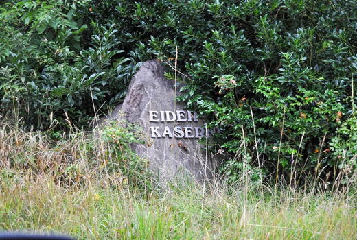 Ein steinerner Wegweiser mit der Aufschrift "Eider Kaserne" steht von üppigem Grün umgeben im Gras.