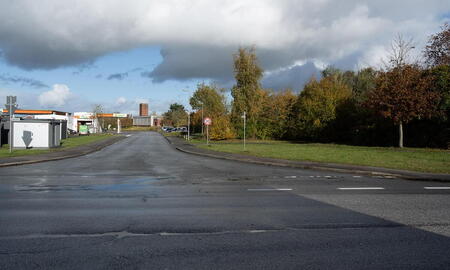 Eine leere Straße führt zum Eingang der Thosberg-Kaserne, gesäumt von Bäumen und Gebäuden unter einem bewölkten Himmel.