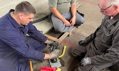 Drei Männer arbeiten gemeinsam in einer Werkstatt an einem mechanischen Projekt auf dem Boden.
