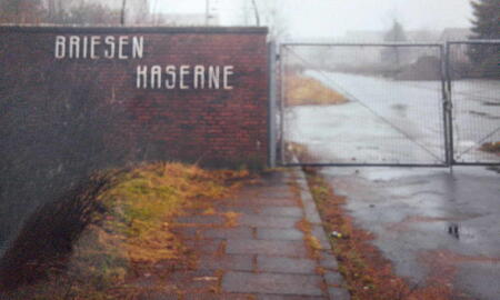 Ein verlassener Kaserneneingang mit einem verrosteten Tor und verblassten Schriftzug auf einer Backsteinmauer an einem nebligen Tag.