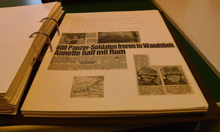 Ein Fotoalbum mit Zeitungsartikeln liegt geöffnet auf einem Tisch, während ein weiteres geschlossen daneben liegt.