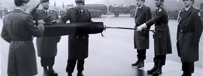 Soldaten in Uniform rollen feierlich eine Fahne ein, während einer von ihnen salutiert.