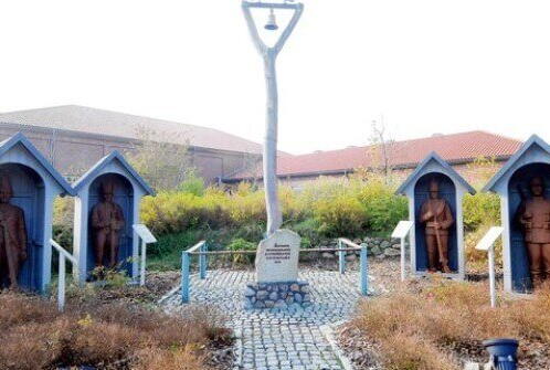 Vier Holzfiguren in kleinen Häuschen umgeben eine zentrale Glocke in einem Garten in Seeth.