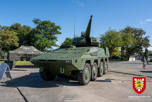 Ein grüner Militärpanzer steht auf einem Freigelände, mit einem getarnten Fahrzeug und Bäumen im Hintergrund.