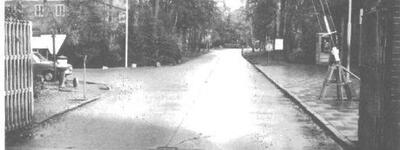Eine leere, schmale Straße führt durch einen bewaldeten Bereich mit einem geöffneten Schranken in einer schwarz-weißen Aufnahme.