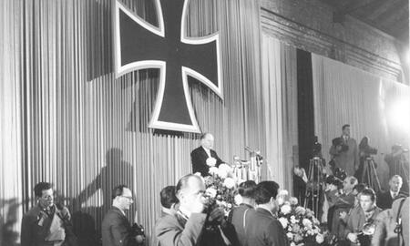 Ein Redner steht auf einer Bühne unter einem großen Kreuz, während Fotografen und Gäste die Ansprache verfolgen.