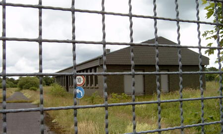 Ein verlassenes Gebäude in Beringstedt ist hinter einem Metallzaun zu sehen, umgeben von überwucherter Vegetation.