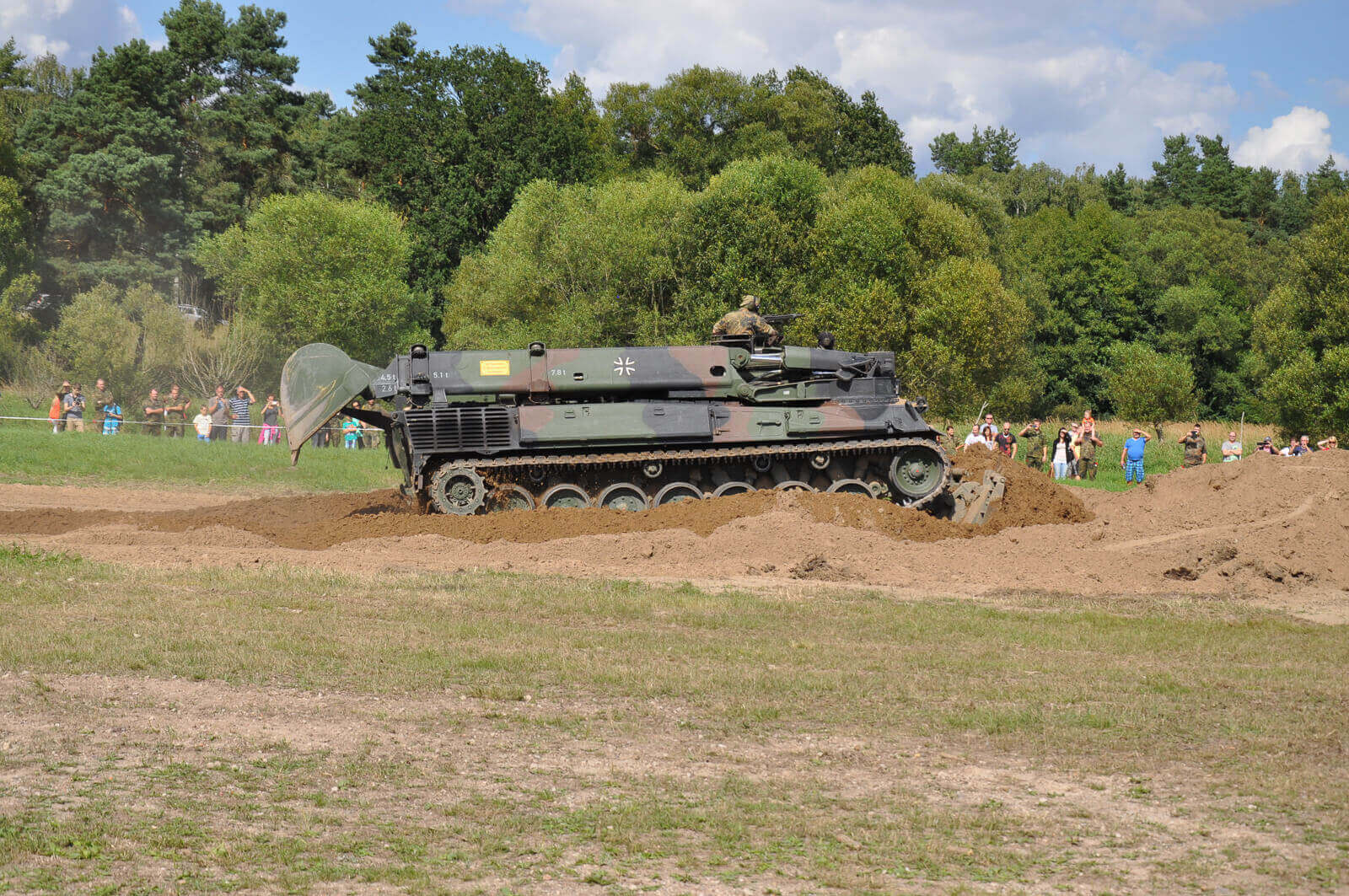 Ein Panzer bewegt sich durch eine sandige Grube, während Zuschauer aus sicherer Entfernung zusehen.